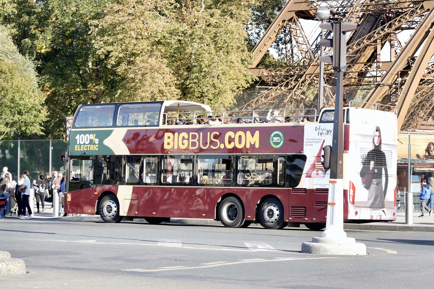 A red Big Bus tourist double-decker stops right next to the steel girders of the Eiffel Tower at a Parisian